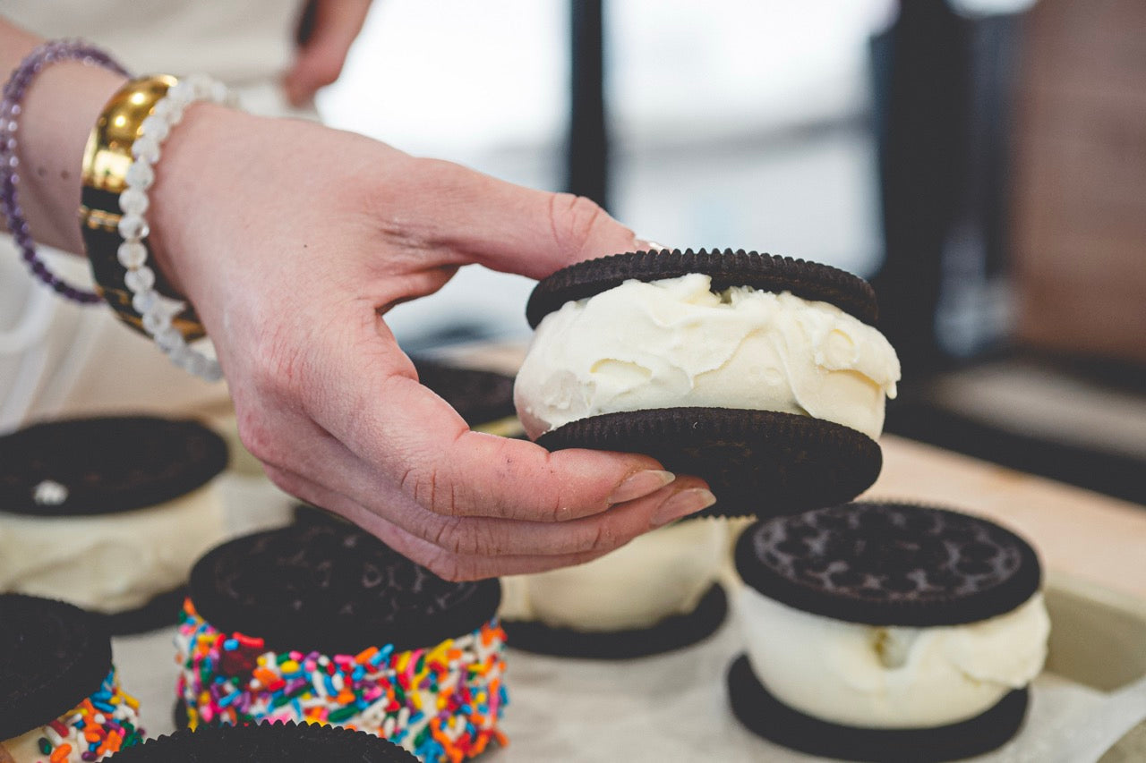 Hand holding a cookie ice cream sandwich with more ice cream sandwiches in the background.