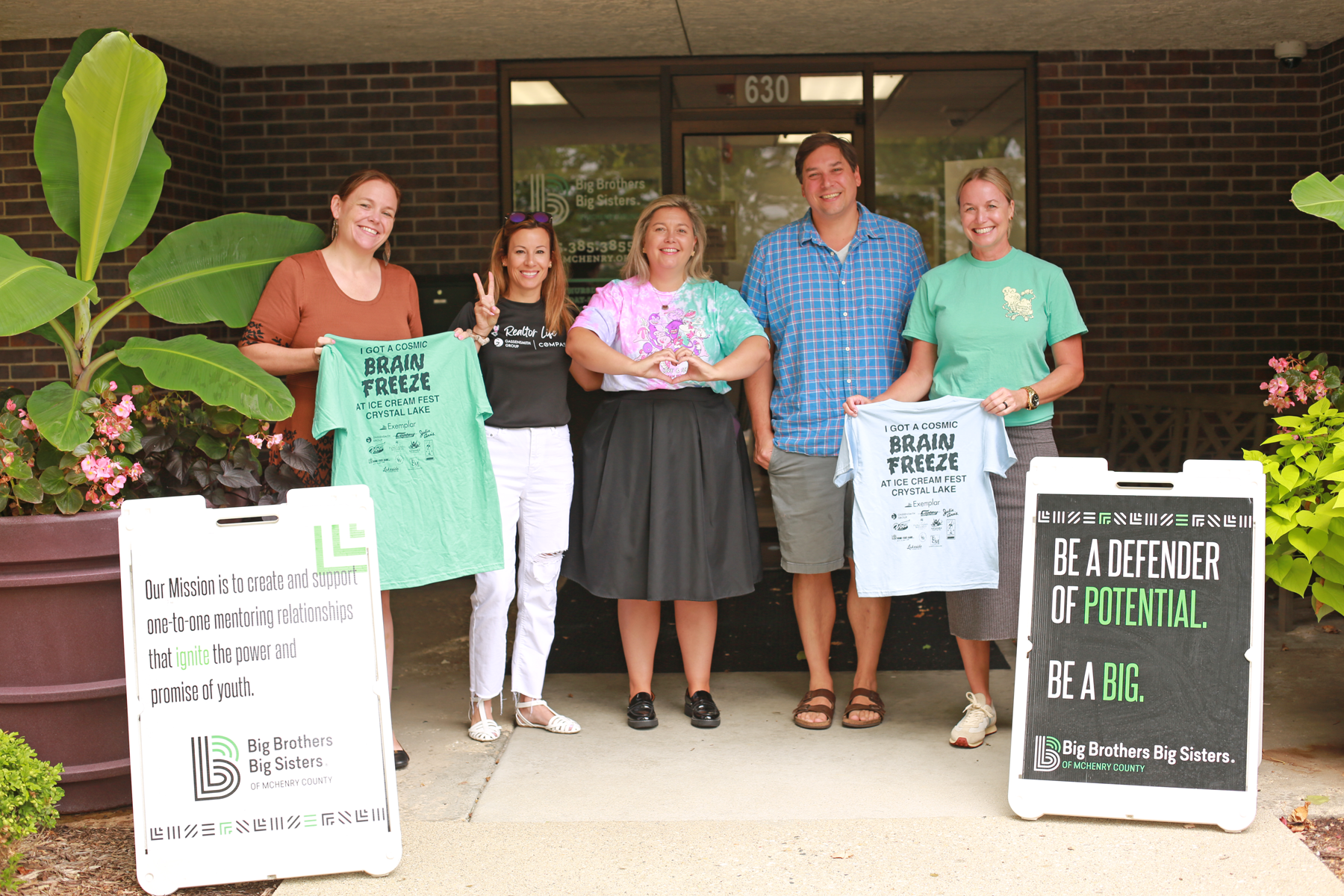 Group of people holding 'Brain Freeze' t-shirts in front of Big Brothers and Big Sisters building with signs.